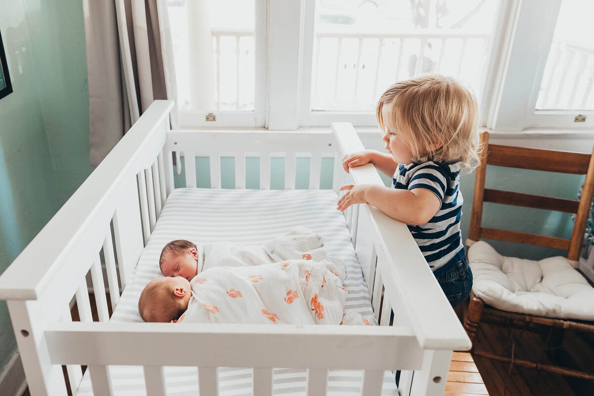 big brother looks over crib rail at his twin newborn baby siblings