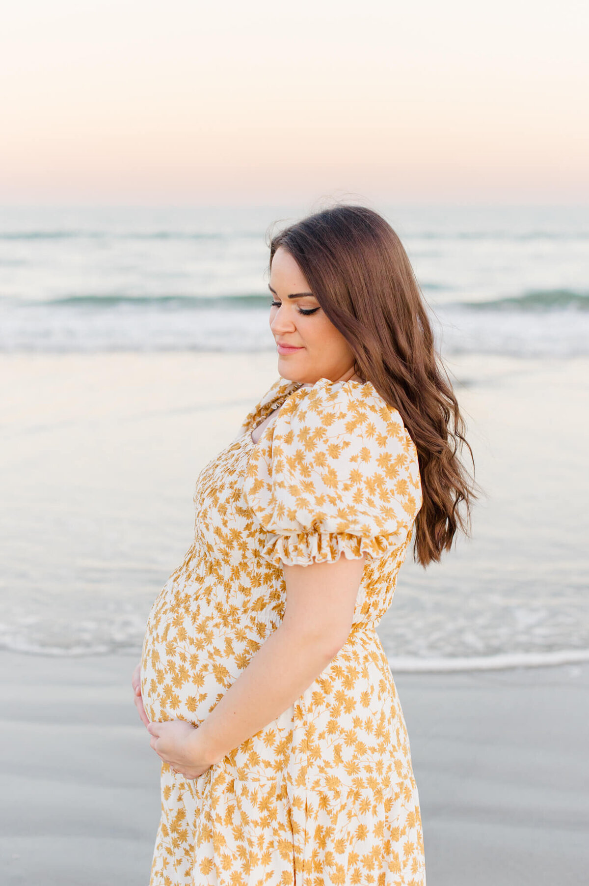 Expectant mother holds her belly and looks down at her baby on the beach a shore drive from Orlando