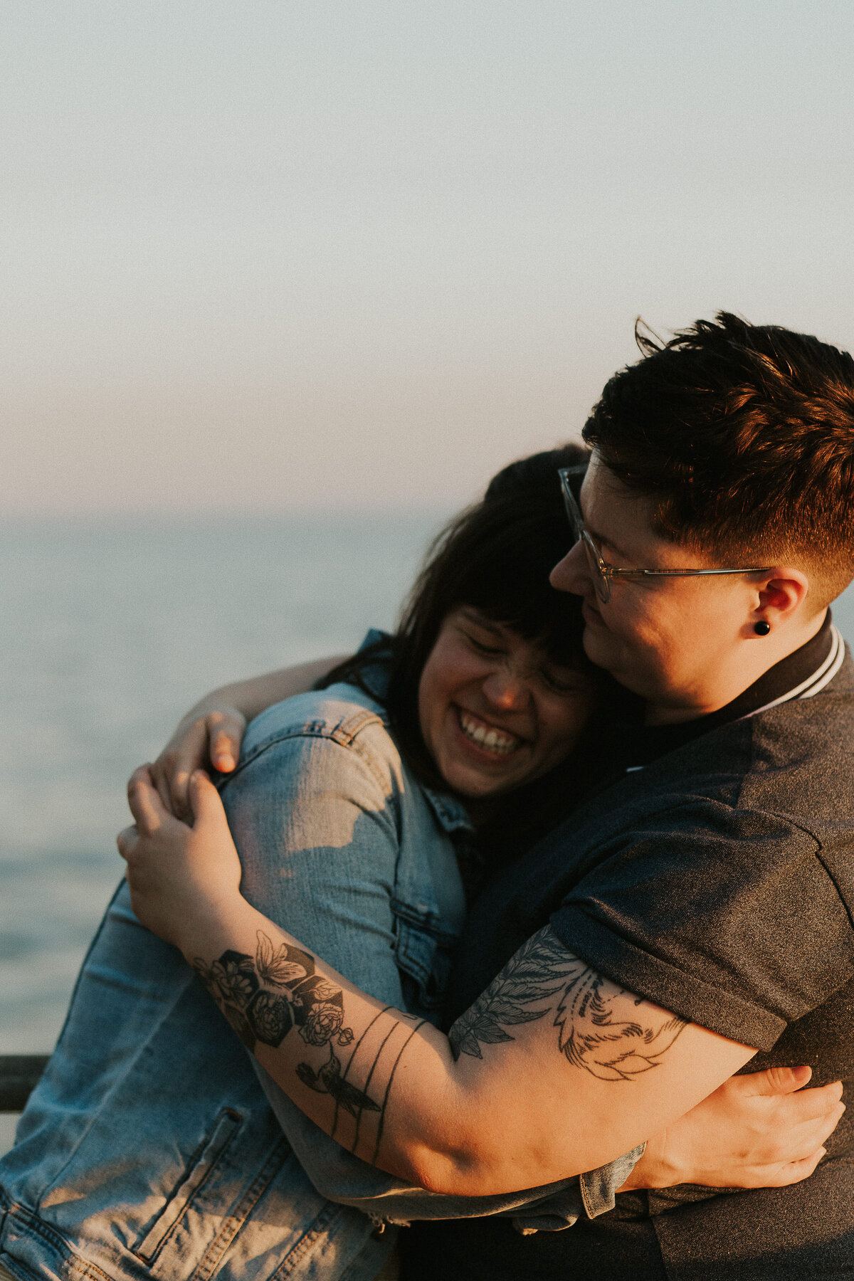 A couple embraces by the water, both smiling brightly as the sun sets.