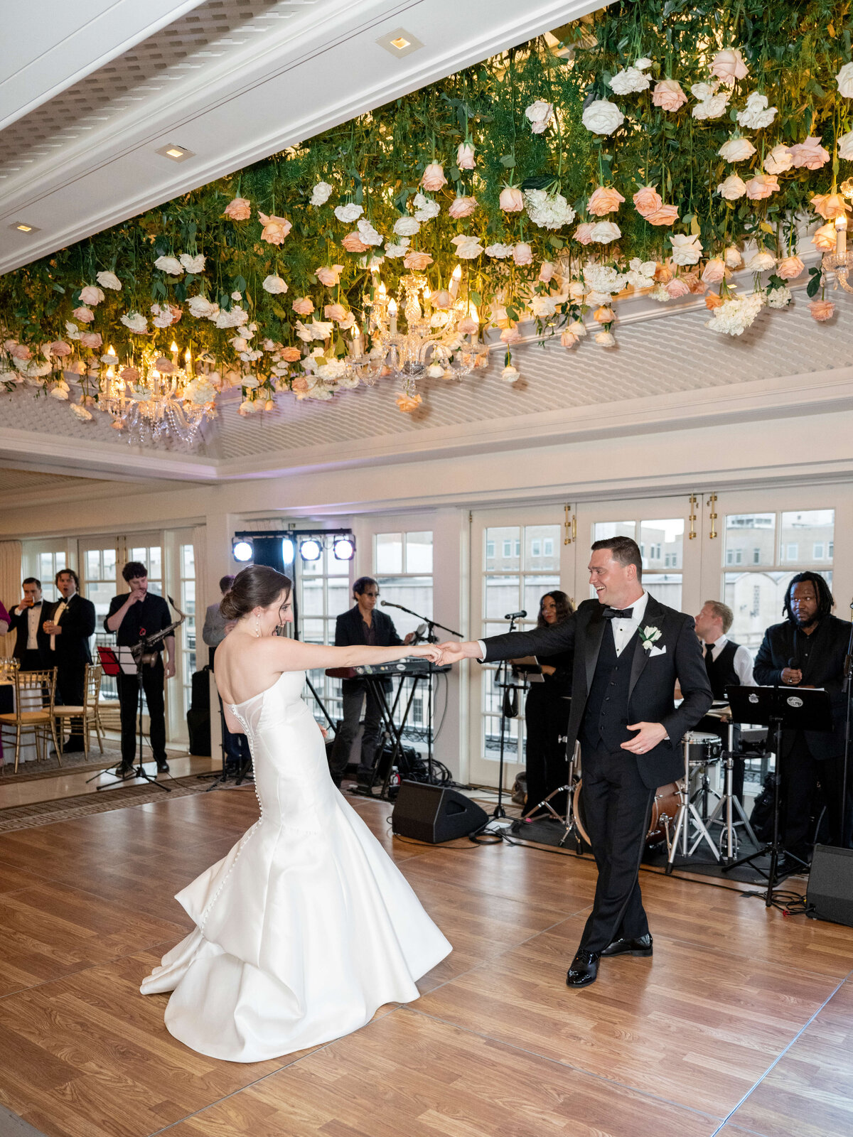 A bride and groom dance in a decorated venue with a flower-covered ceiling. The bride wears a strapless white gown, and the groom is in a black tuxedo. A live band plays in the background, and guests look on.