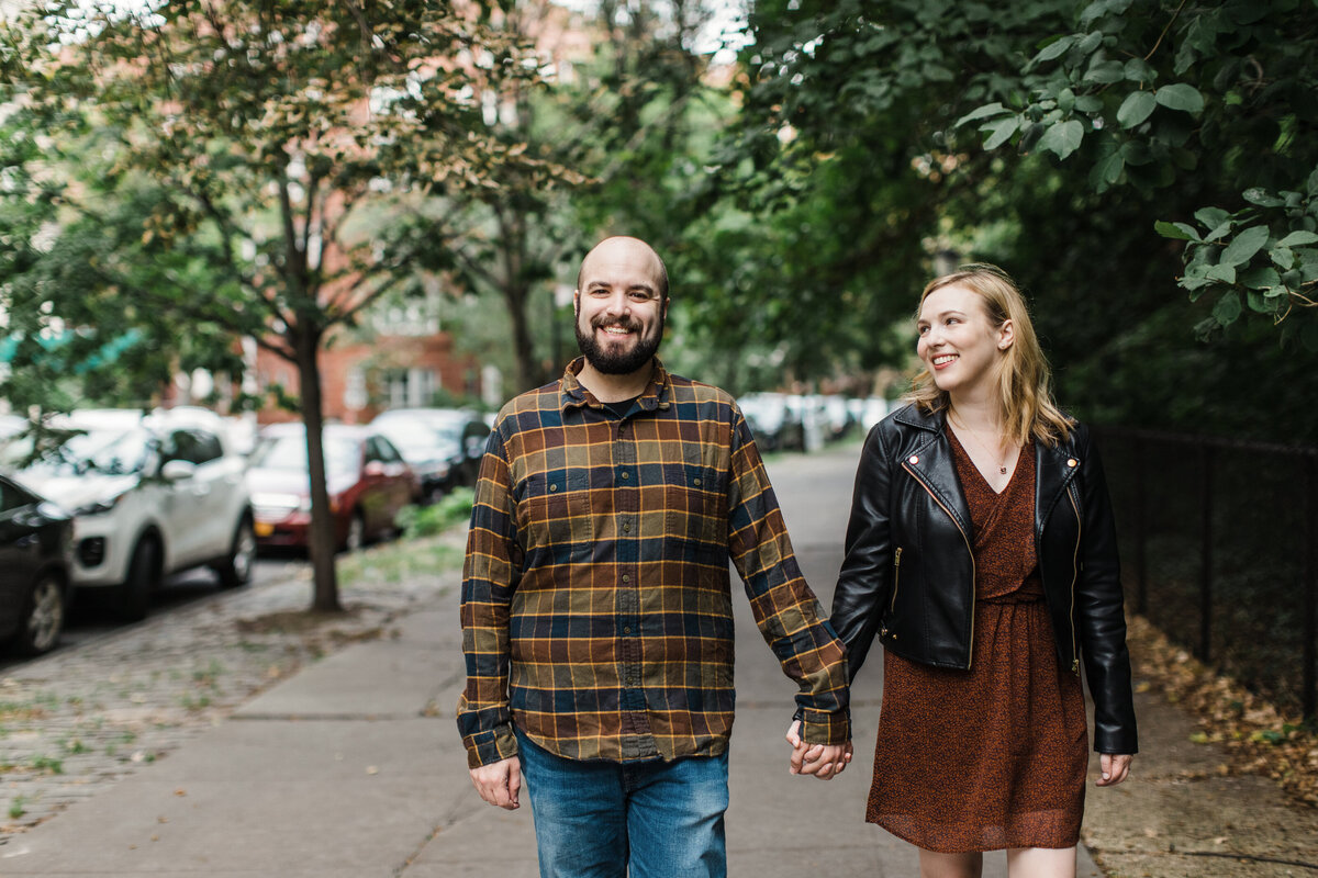 A couple holding hands and walking down the sidewalk during their engagement session in Park Slope in Brooklyn, New York. The woman on the right is wearing a detailed, short, dark, orange dress with a black leather jacket. The man on the left is wearing a flannel shirt and jeans.