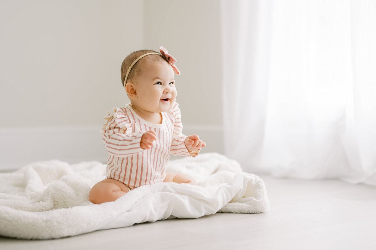 Baby girl in pink and white striped romper with pink bow smiles sitting on white blanket in Raleigh NC photography studio