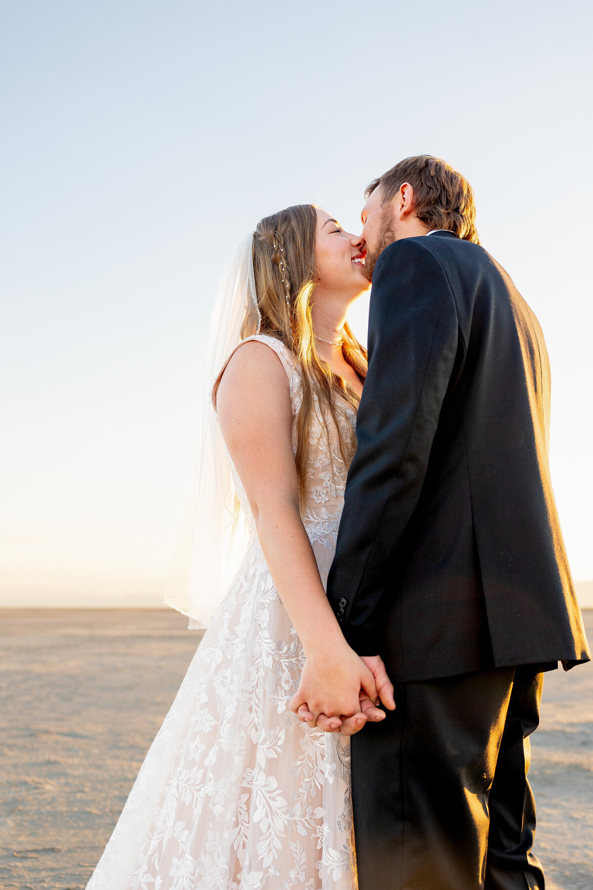 Bride and groom share a kiss while holding hands, glowing in the golden morning light.