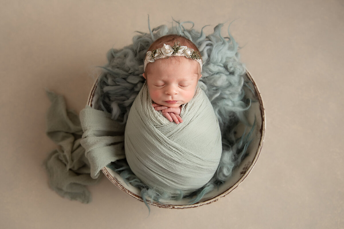 Newborn baby wrapped in a seagreen wrap lying on matching fur in a wooden bowl on a  cream background.