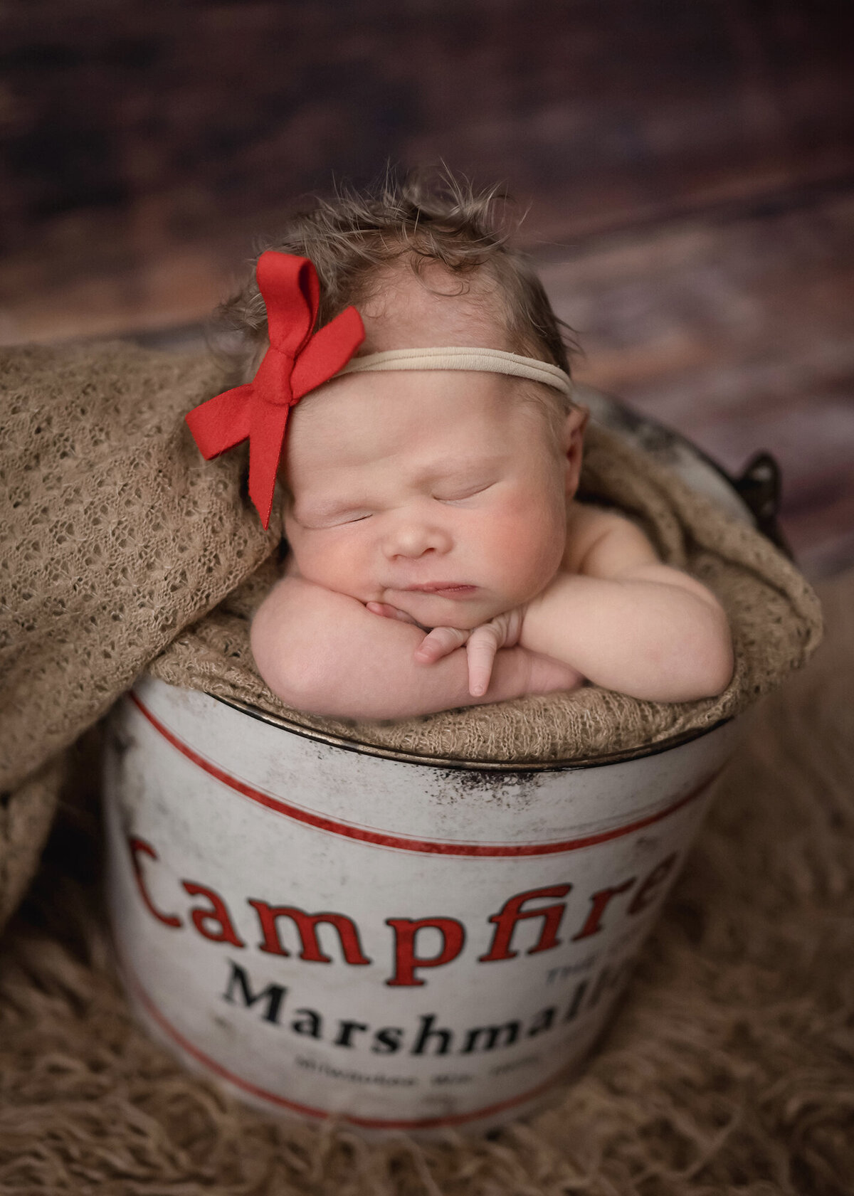 A baby sleeps squished on her arm in a red bow inside a metal campfire bucket with brown blanket