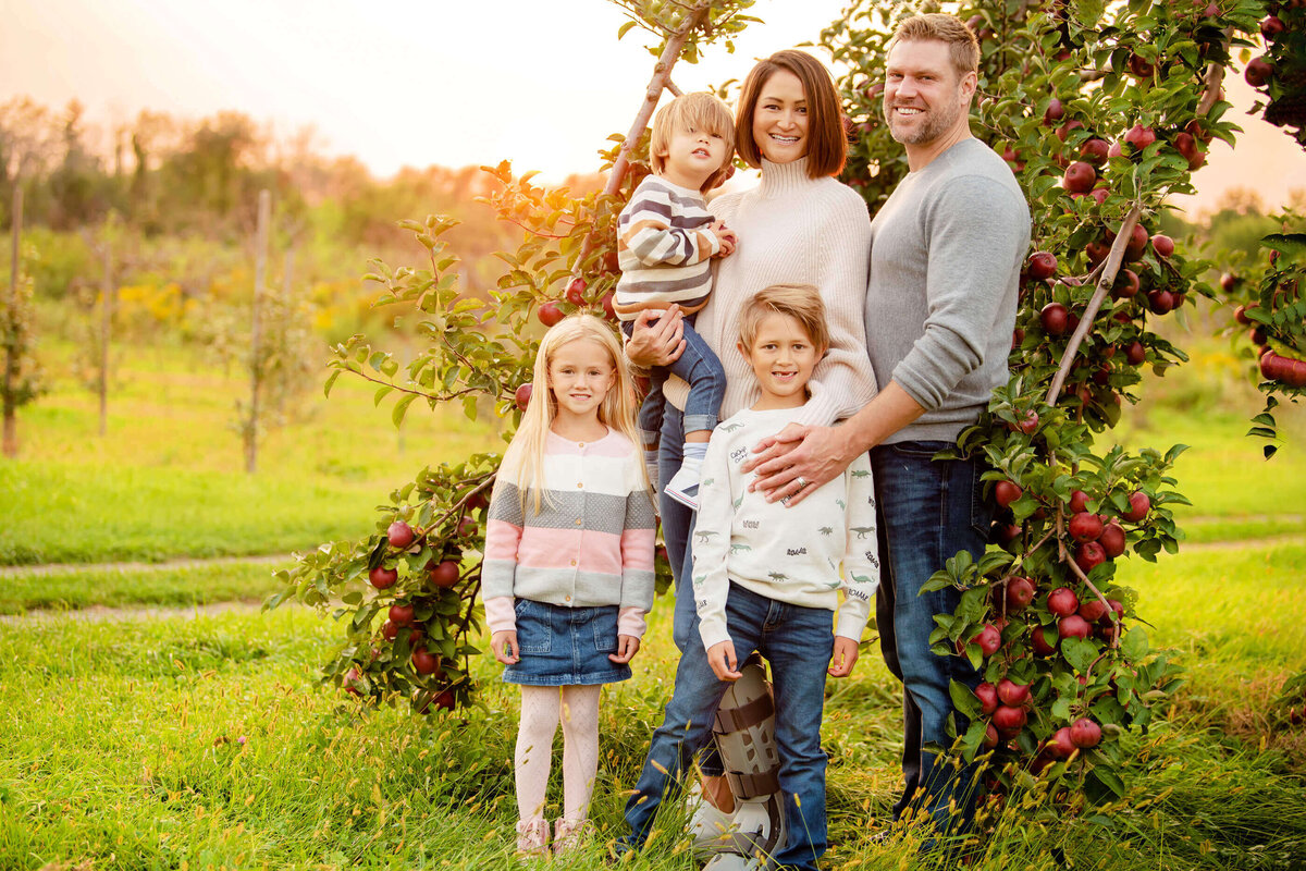 mom and dad with their three kids standing near an apple tree in the golden hour for their fall family photos in Burlington, ON