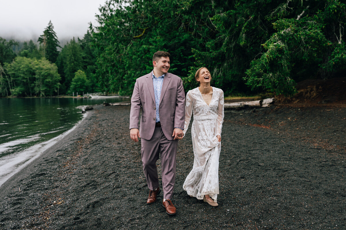 A Puerto Rican bride wearing a cream shawl and holding dried flowers laughs as she looks at her groom in a gray suit jacket and blue polka dot shirt as snow falls among the Evergreen trees at their Columbia River Gorge PNW winter adventure elopement at Government Cove near Hood River, Oregon. | Erica Swantek Photography