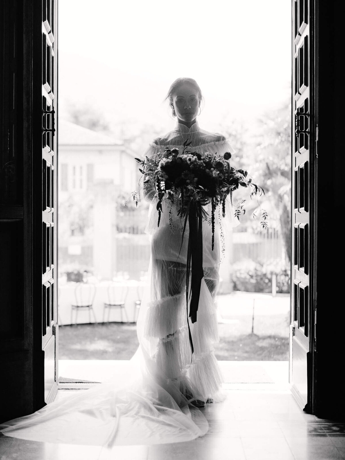 Bride holding a flower bouquet standing in the middle of a huge door on a blurred white bright light background