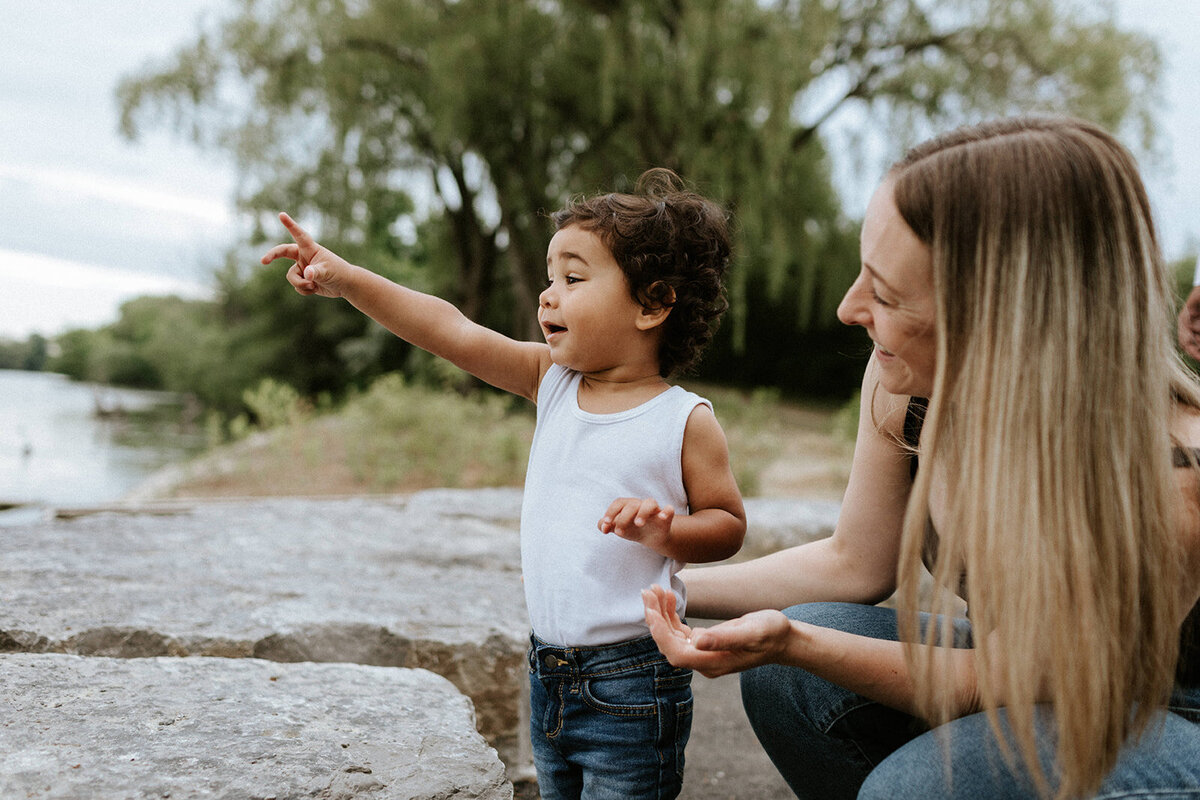 A child in a white tank top points toward the water, standing beside a woman with long hair. Captured by a Toronto photographer, they are outdoors near a lake, surrounded by lush trees.