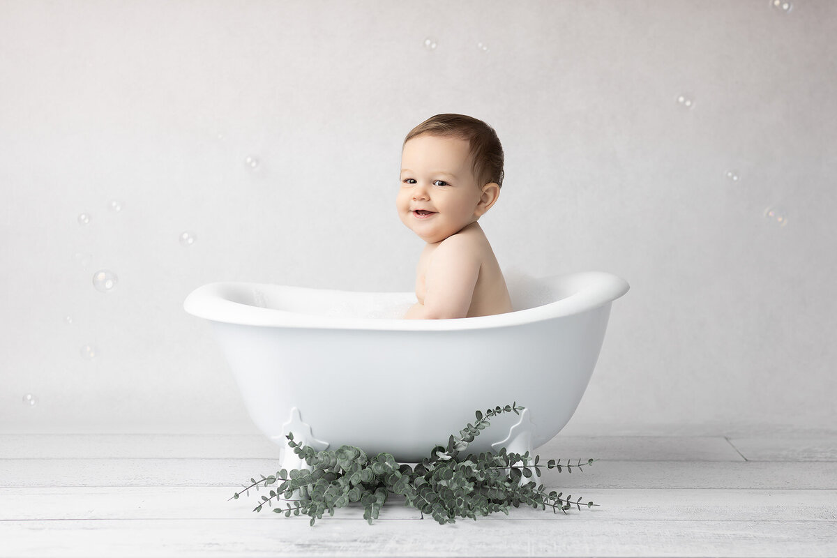 Smiling baby sits in a vintage white bathtub surrounded by bubbles and eucalyptus during a DFW milestone photography session.