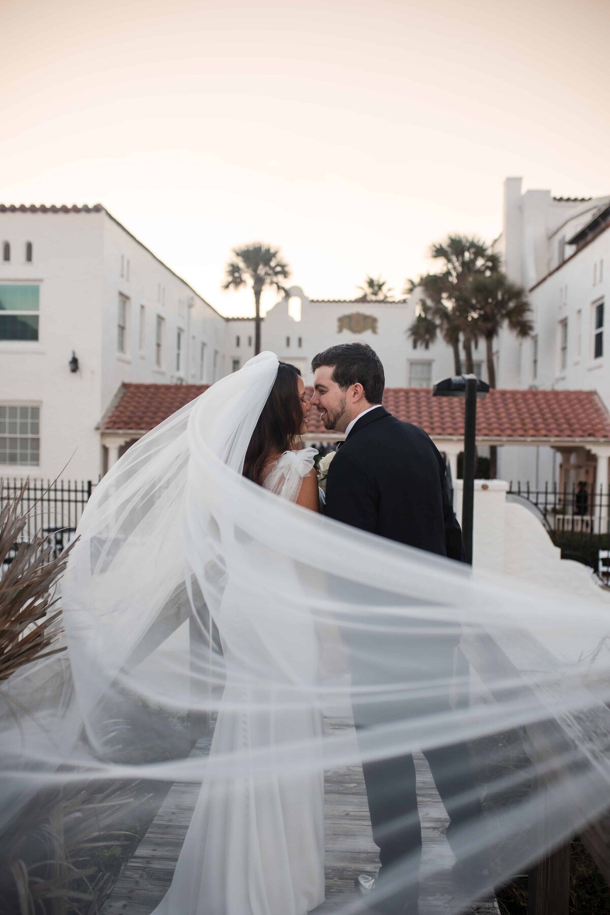 A romantic post-ceremony moment between a bride and groom at Casa Marina in Jacksonville Beach, Florida. The couple shares a kiss as the bride’s cathedral-length veil catches the breeze, flowing gracefully in the foreground. Surrounded by the historic white architecture and palm trees of the venue, the scene captures a timeless coastal elegance. Photographed by Phavy Photography in a true-to-color, documentary style that highlights movement, emotion, and the beauty of golden hour.