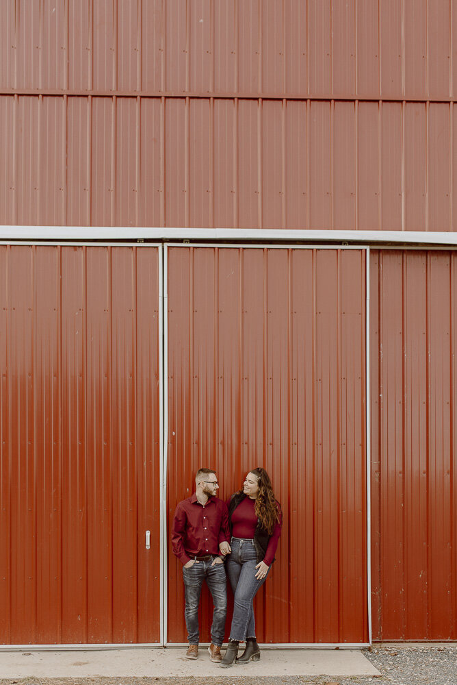 Couple stands in front of red barn while smiling at each other