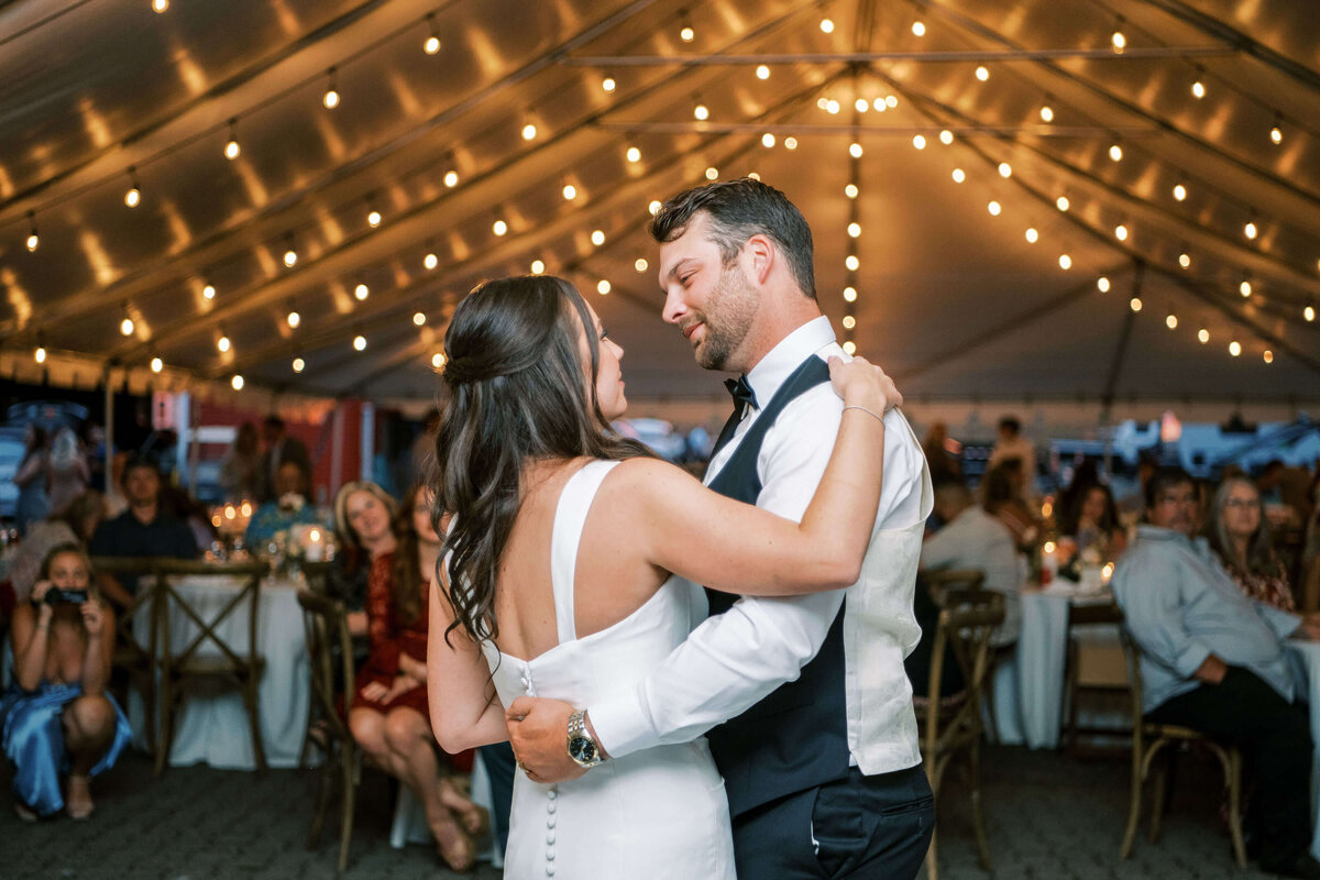 A bride and groom have their first dance, in the background you can see the outdoor tent lit up with string lights.