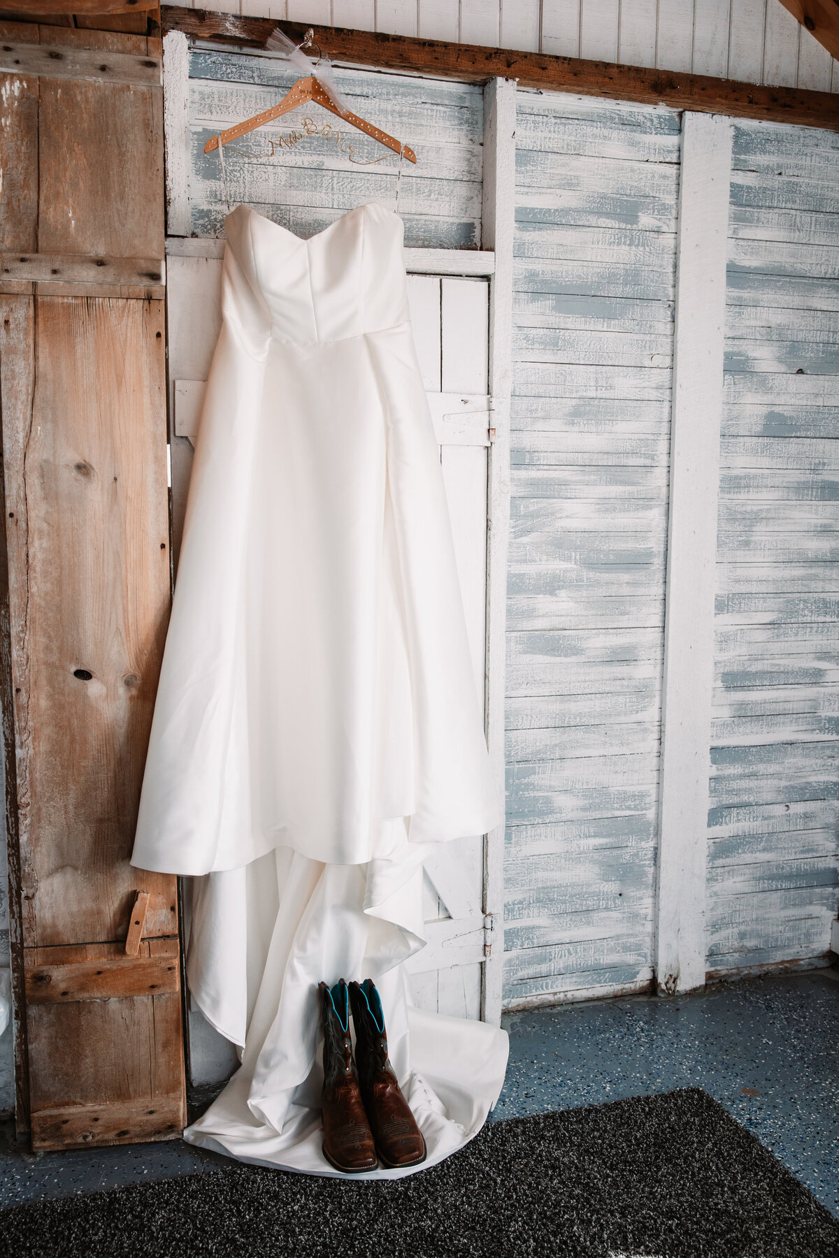 A bride's dress hangs on a rustic wall and doorframe with her boots below it