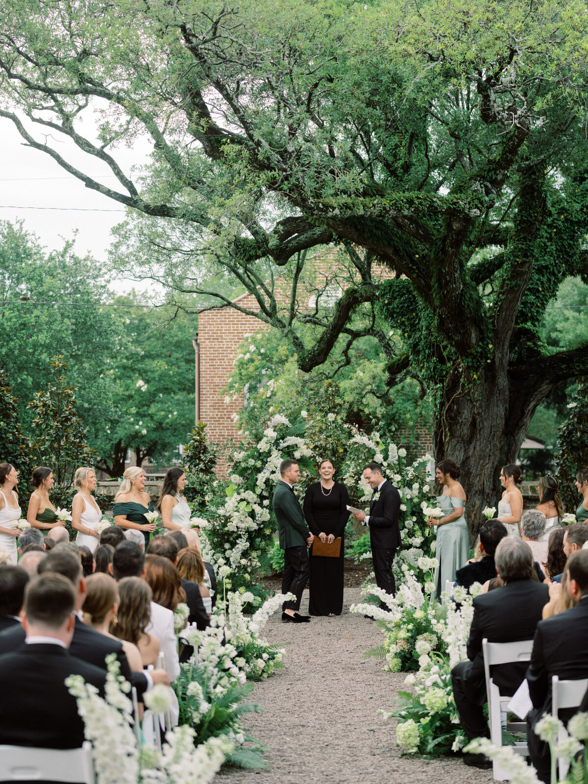 This enchanting outdoor wedding scene in Charleston captures joyful couples exchanging vows beneath a lush tree, surrounded by beautiful floral arrangements and attentive guests.