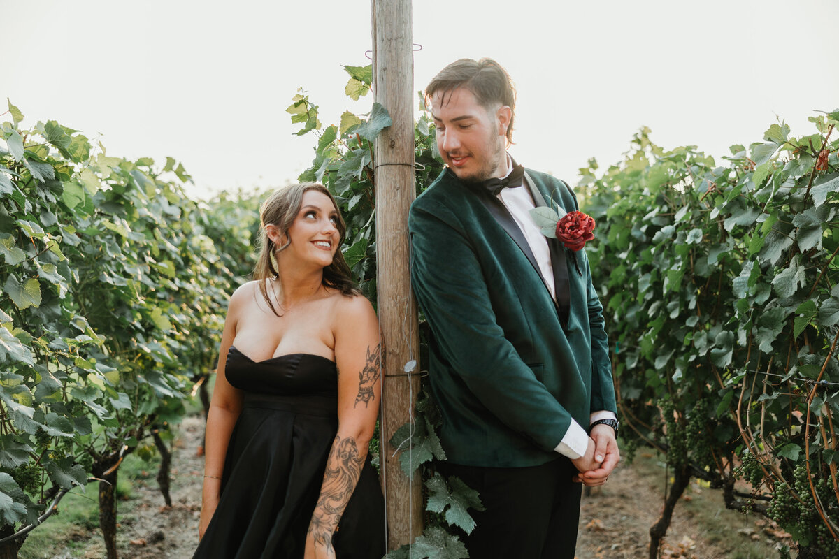 A bride in a black wedding dress and her groom smile at each other as they turn to look at each other in a vineyard