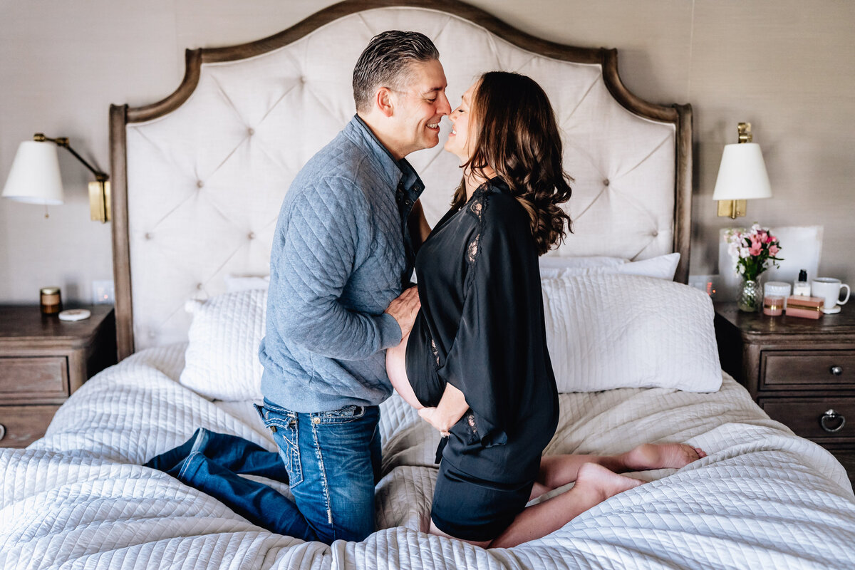 A couple kneels on a bed, leaning in for a tender kiss. The pregnant woman wears a black robe that drapes over her belly, and the man lovingly holds her waist