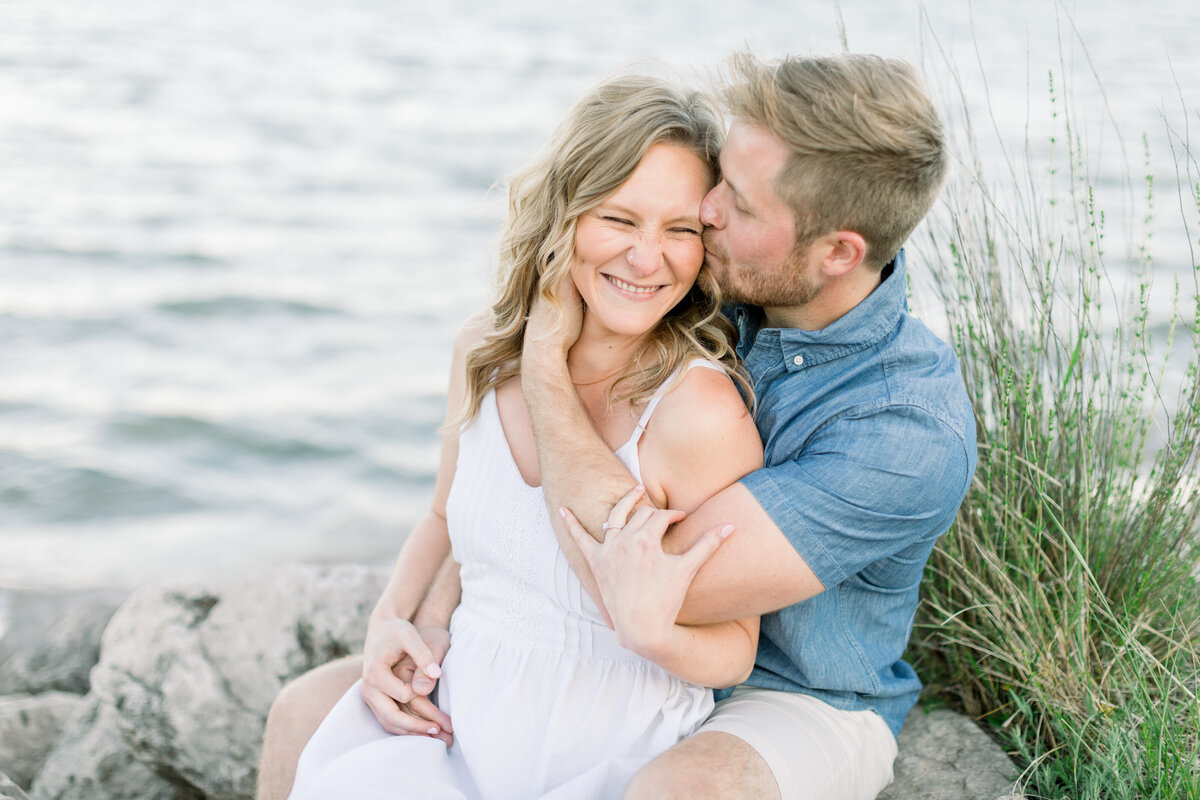 Close up of a woman smiling and laughing while her fiance kisses her face by Lake Michigan in Sheboygan, WI