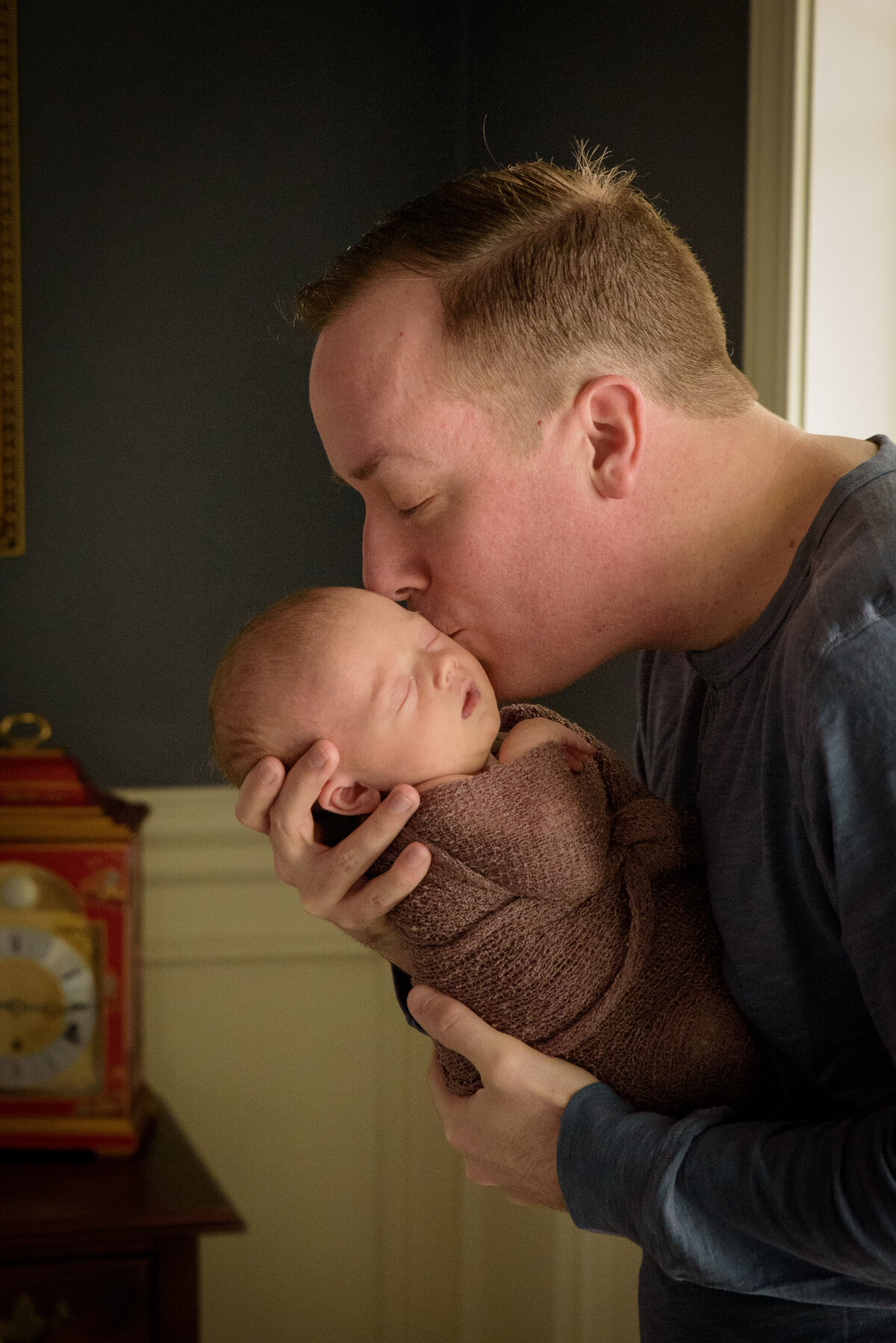 Dad is holding and kissing newborn daughter who is wearing a dusty rose wrap in front of a window in their home in Green Bay, Wisconsin