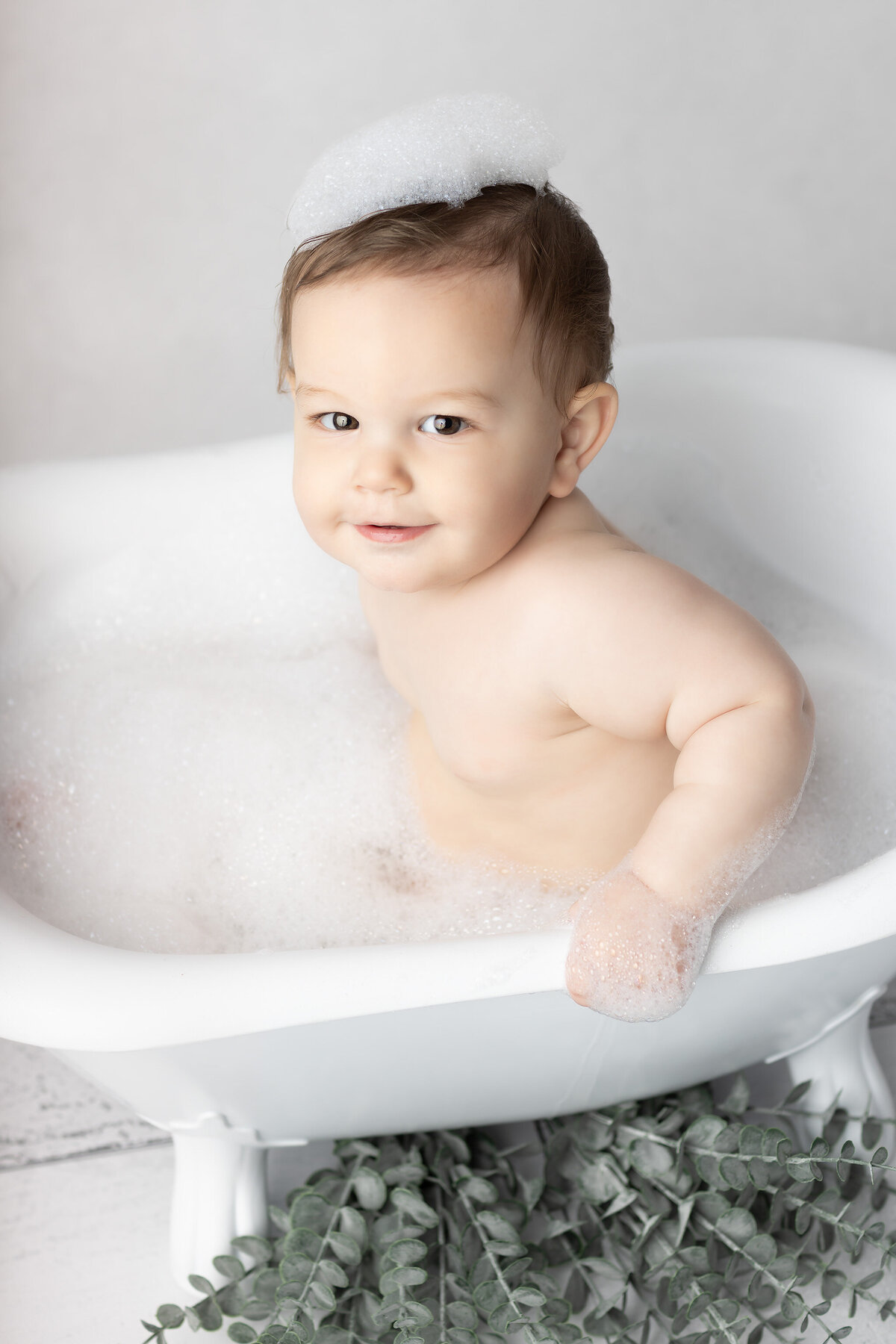 DFW milestone photography session with a baby smiling in a vintage-style white tub, topped with playful bubbles on their head and surrounded by soft greenery.