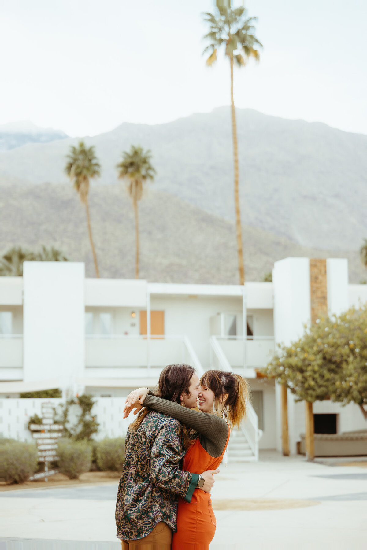 Couples photography session at Ace Hotel in Palm Springs. Woman wears orange overalls and man is in paisley teal shirt.  White hotel and palm trees in background.
