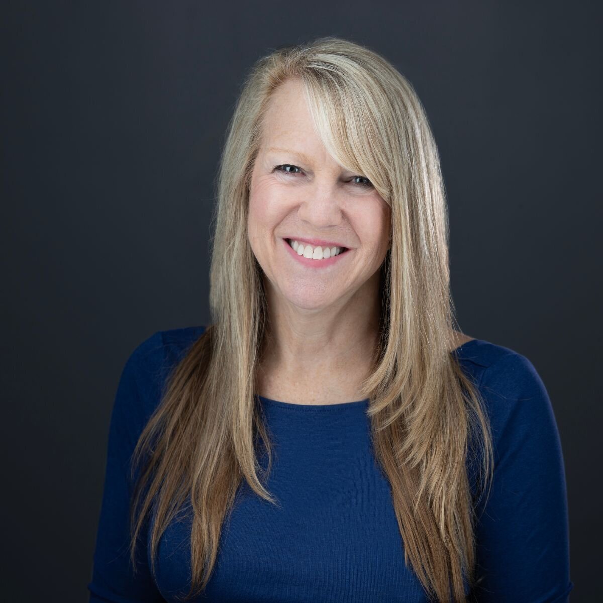 A headshot with a dark grey background of a woman with blond hair and smiling big at the camera. She is wearing a blue shirt and projecting confidence and approachability. Photo by sacramento headshot photographer, philippe studio pro.