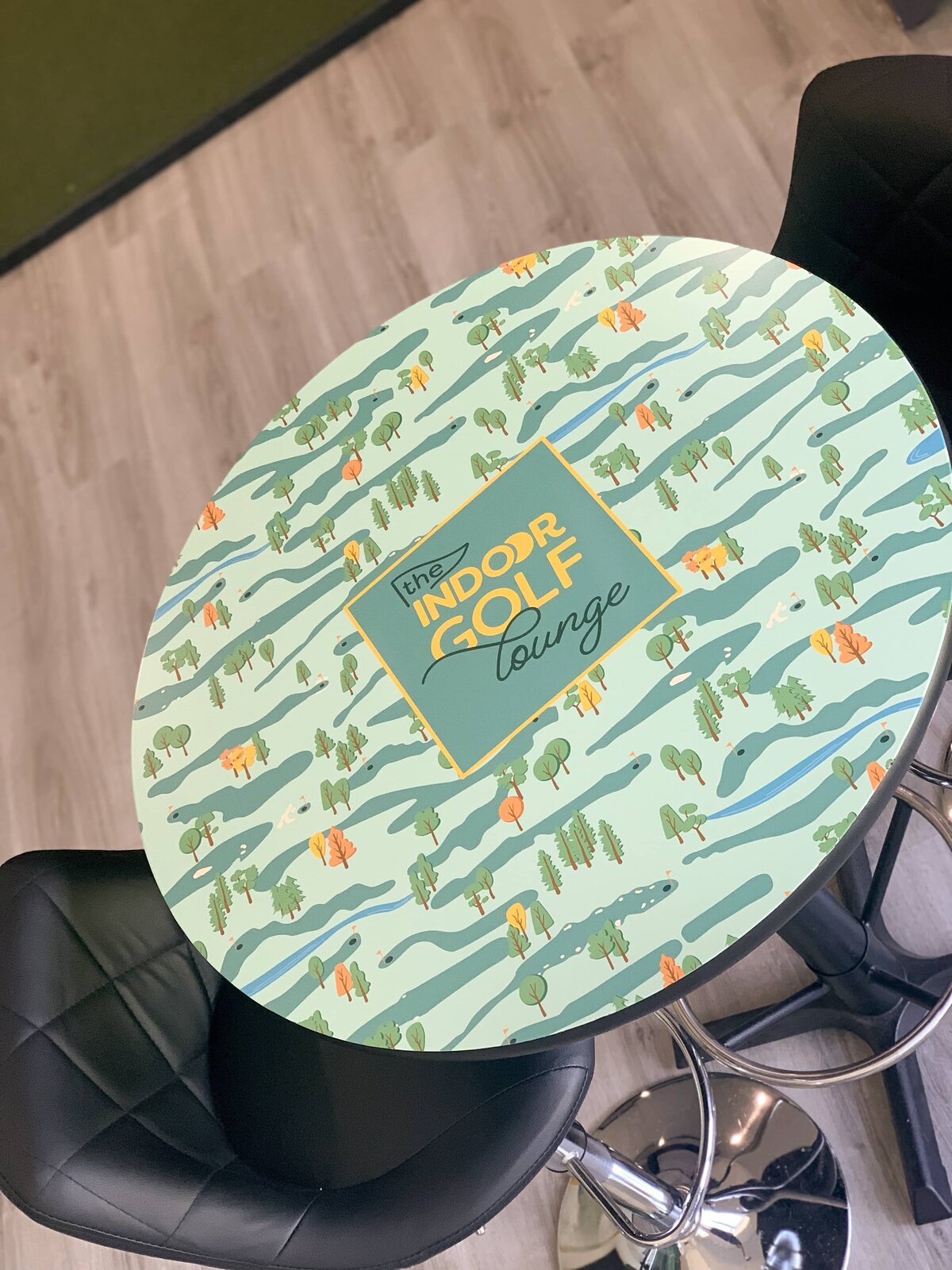 A round, high-top table with a custom printed top. The Indoor Golf Lounge brand pattern with a light green background with the logo in the center.