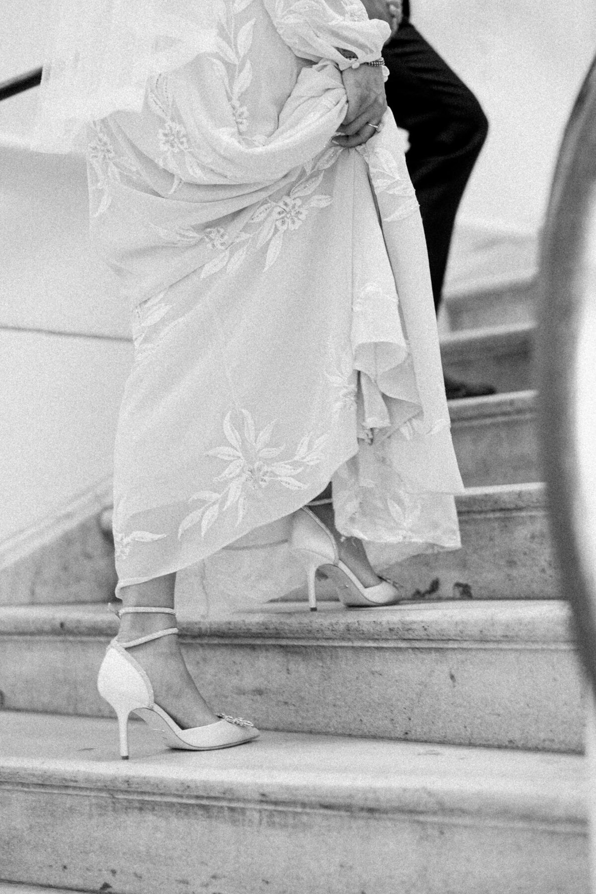 A bride lifting her dress as she ascends stairs with her partner, showcasing her white heels.