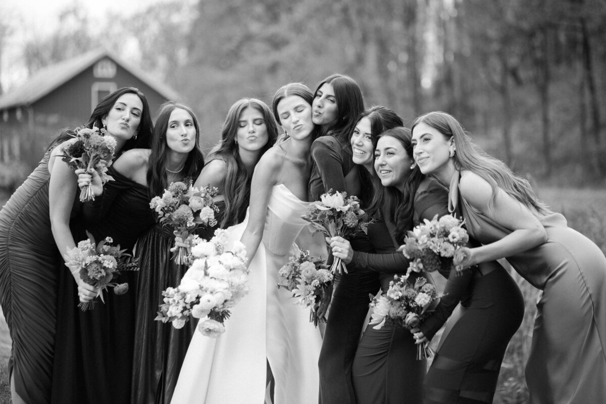 The bride stands surrounded by her bridesmaids, all in elegant dresses, holding bouquets and making happy faces