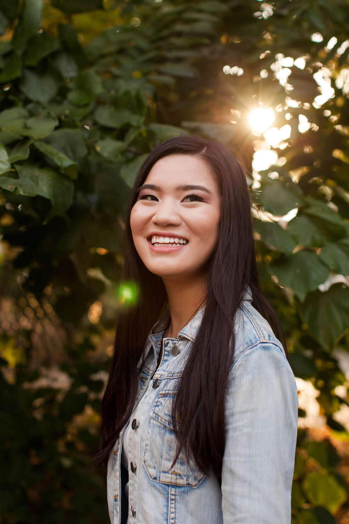 A beautiful black haired teen girl wearing a jean jacket smiles in her senior portrait with sunflares in the bakcground.  Captured by Springfield, MO senior photographer Dynae Levingston.
