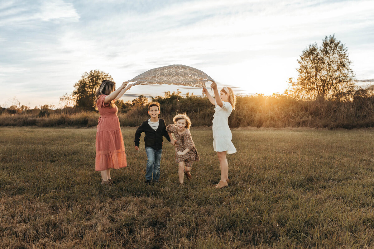 four siblings play red rover under a lace blanket in a field.