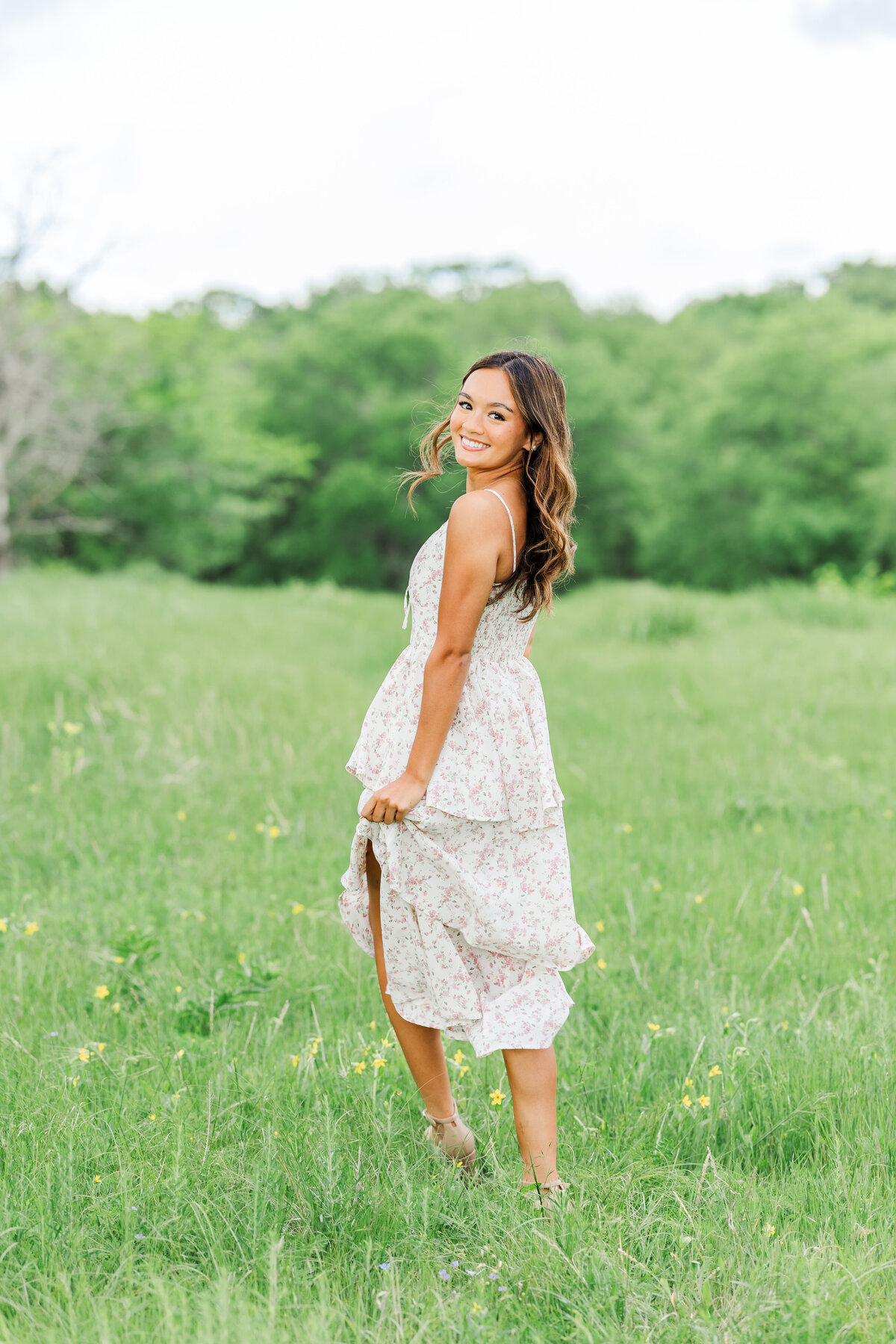 Senior girl smiling over her shoulder in a flowy floral dress, standing in a green field at Arbor Hills Nature Preserve in Plano, TX.
