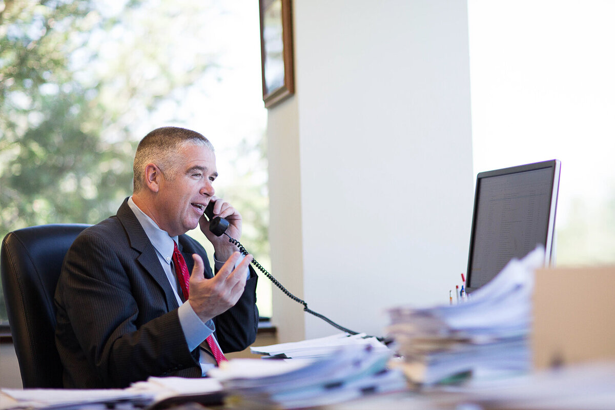 older man in a suit talking on the phone, sitting at a desk, gestuing with his hand