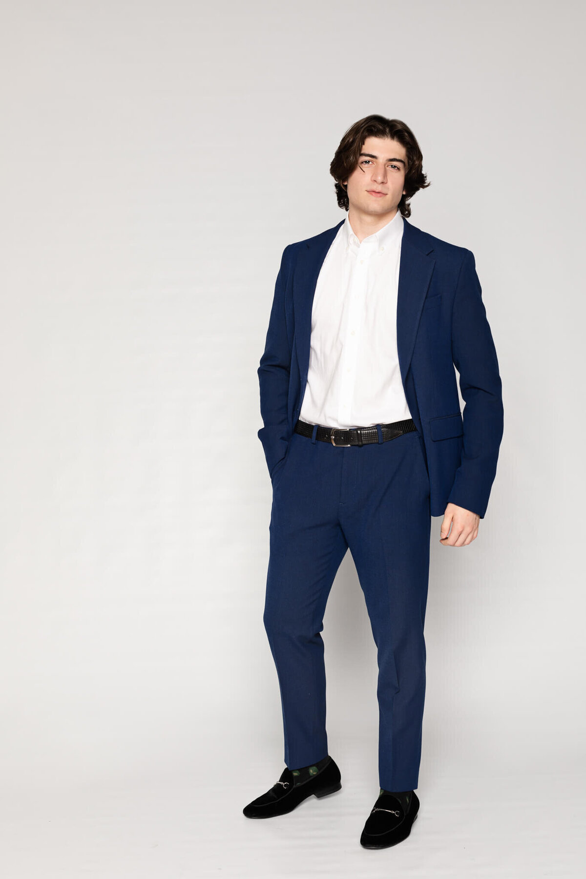 A person with shoulder-length hair is wearing a blue suit and white dress shirt, standing with hands in pockets against a plain white background. They are looking at the camera with a neutral expression.