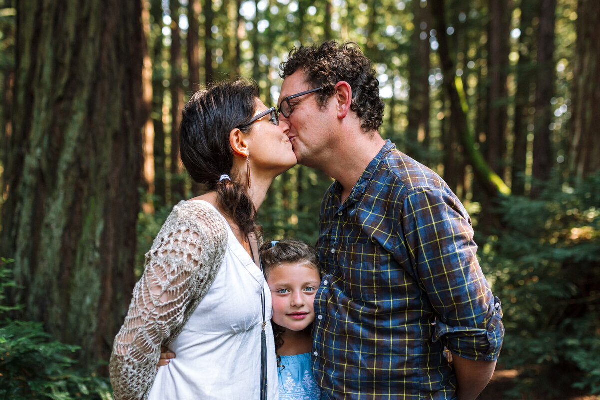 parents standing in Oakland park kissing with 8-year-old daughter standing between them and looking at the camera
