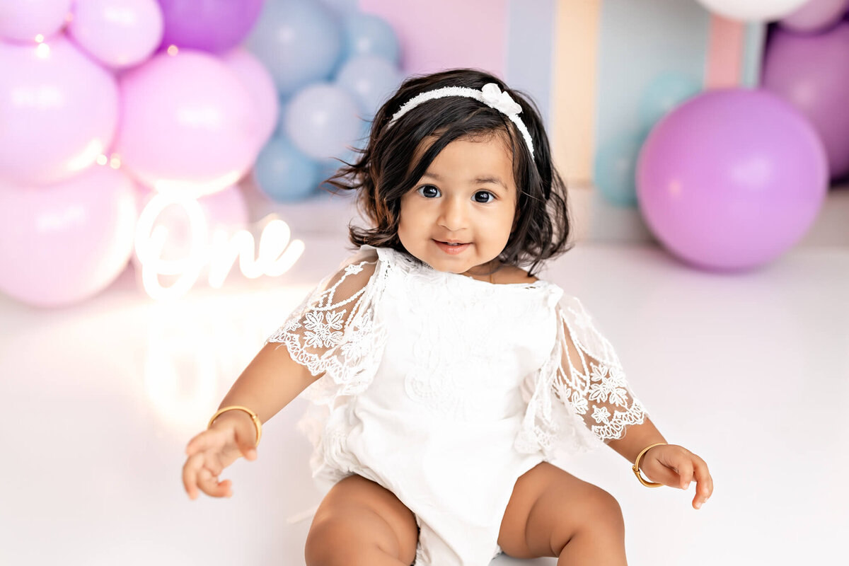 A toddler with dark hair, wearing a white lace dress and headband, sits on the floor. Balloons in pastel shades of pink, purple, and blue surround her. A lit sign reading "One" is in the background.