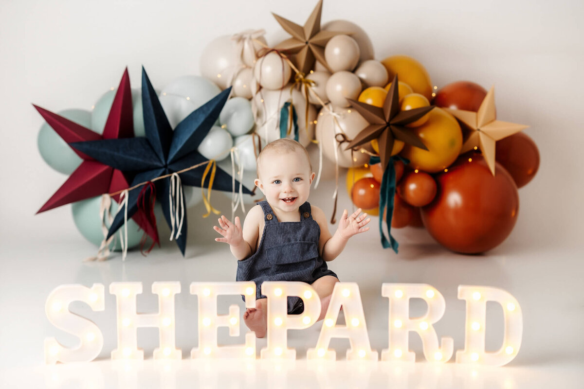 A smiling baby in a dark blue outfit sits surrounded by colorful balloons and stars, perfectly capturing the essence of joyful families photography. The name "SHEPARD" is displayed in illuminated letters against the white backdrop, showcasing a portrait photographer's skill.