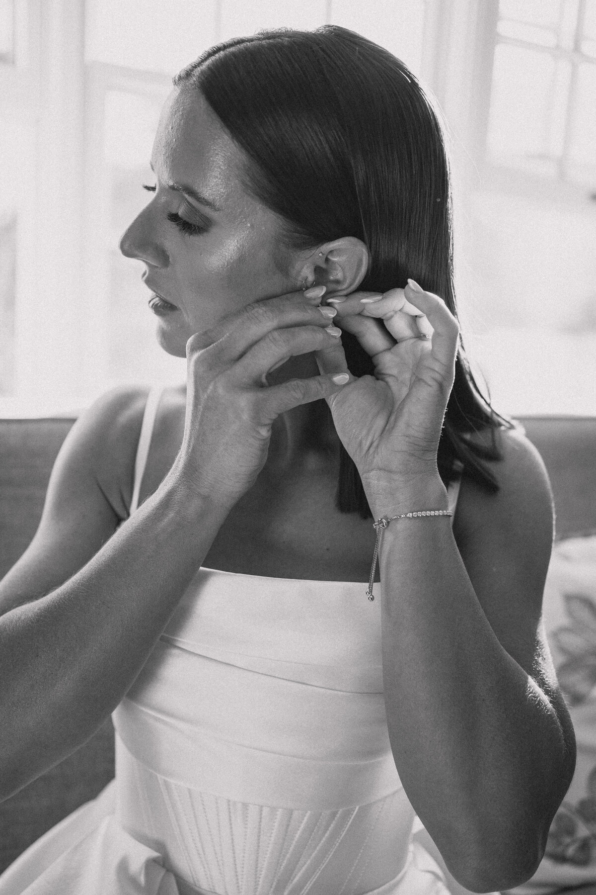 Bride adjusting her earring while getting ready for her wedding, captured in an elegant black-and-white photograph.