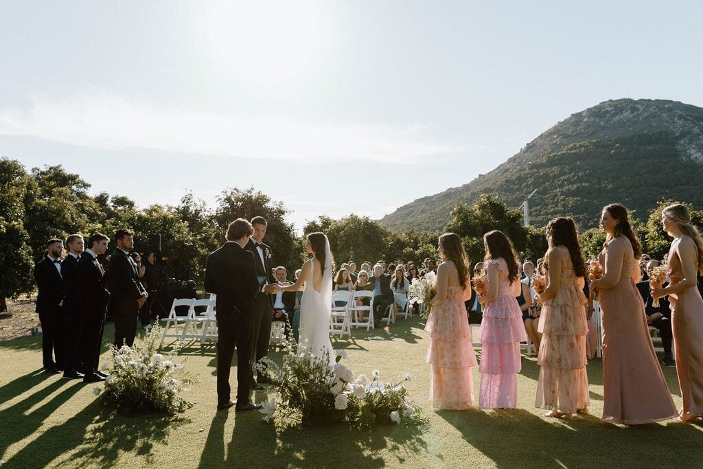 Bride and groom being married outside surrounded by trees and hills. Guests in the background, groomsmen and bridesmaids on either side of the new couple 