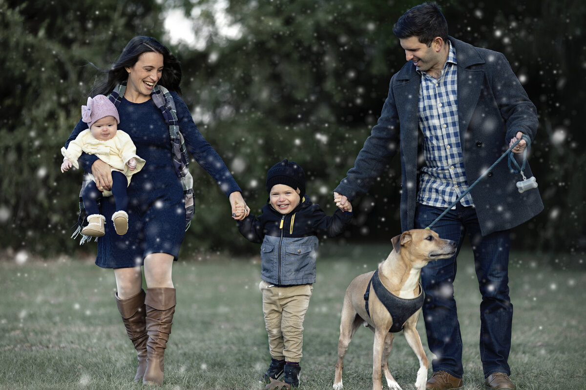 A mom and dad walk through falling snow in a park with their infant daughter in mom's arm and holding hands with their toddler son while walking the dog