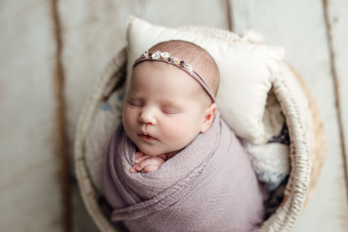 Studio newborn photography - baby girl sleeping in woven basket on top of distressed painted hardwood floor. Baby is wrapped in pale purple wrap and coordinating delicate floral headband.