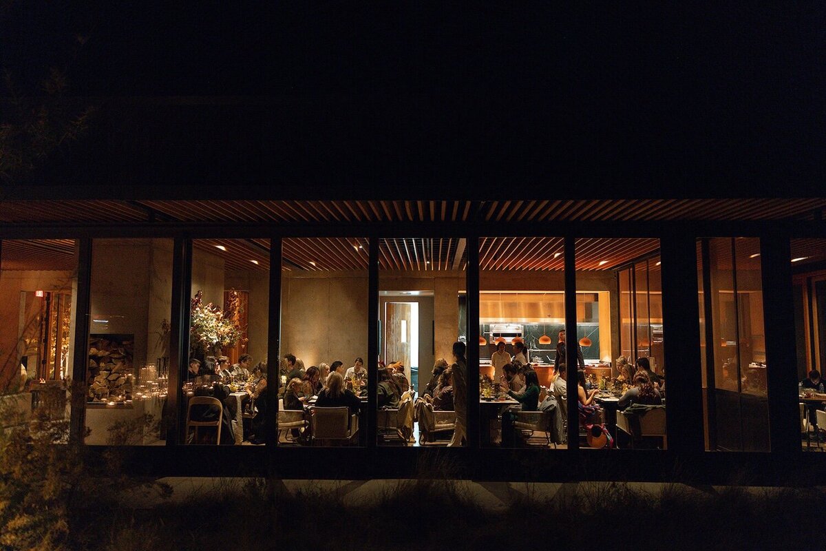 A moody and romantic  rehearsal dinner at Camp Sarika at Amangiri Resort in Utah. Guests eat dinner inside a cozy modern room with glowing candles.