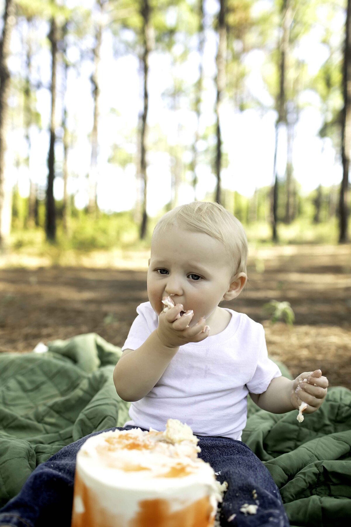 Birthday boy in suspenders and overalls, with cake frosting on his hands, having fun smashing his cake under the shade of pine trees