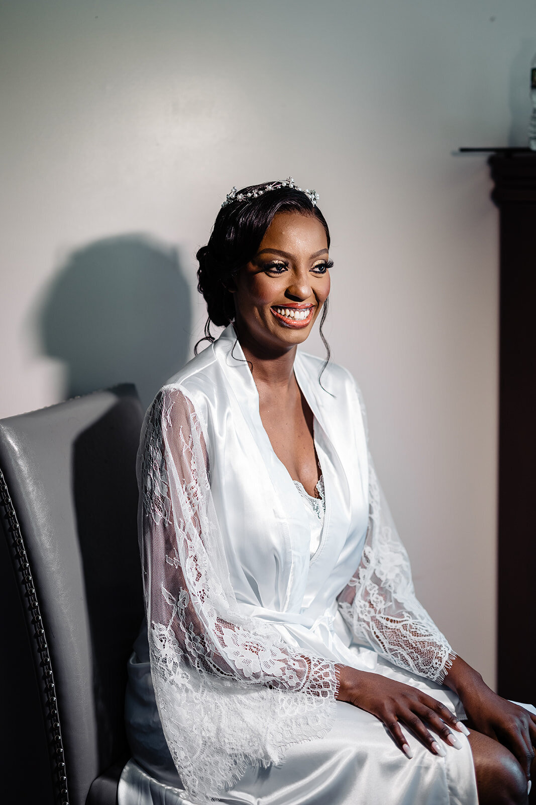A radiant bride sitting in a well-lit room, smiling gently, wearing a white dressing gown with lace detailing and a delicate tiara, exuding happiness and elegance before the wedding ceremony.