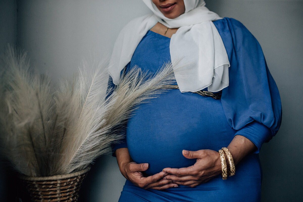 Maternity photoshoot with woman wearing blue dress, white scarf, and holding her belly.
