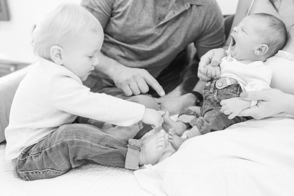 Black and White photo of a toddler counting the toes of his neborn baby brother during an in-home session by Virginia Beach NEwborn Photographer Mary Eleanor