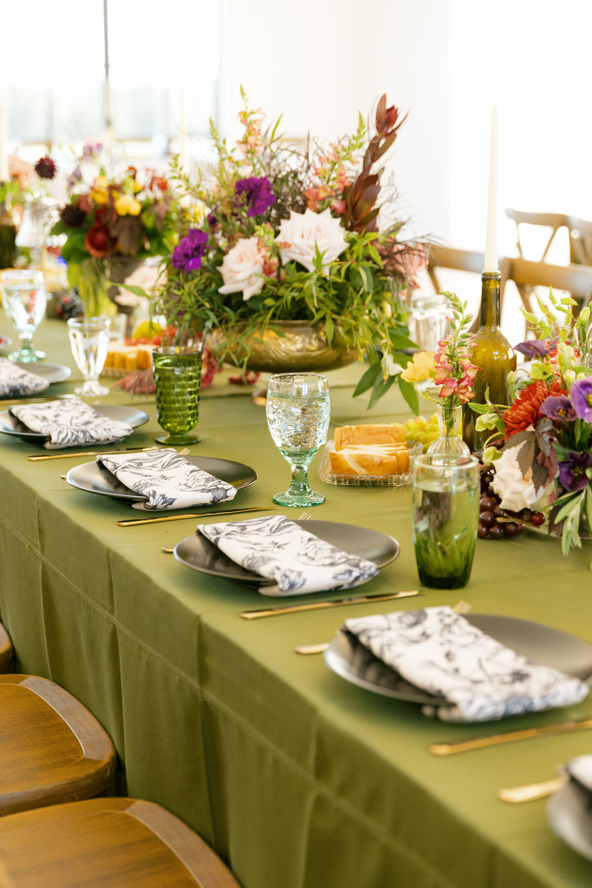 A reception table design with a dramatic garden-style floral centerpiece featuring roses, dahlias, greenery, and deep-toned blooms, surrounded by candles, fruit details, and eclectic vintage glassware, created by a wedding florist for a refined celebration.