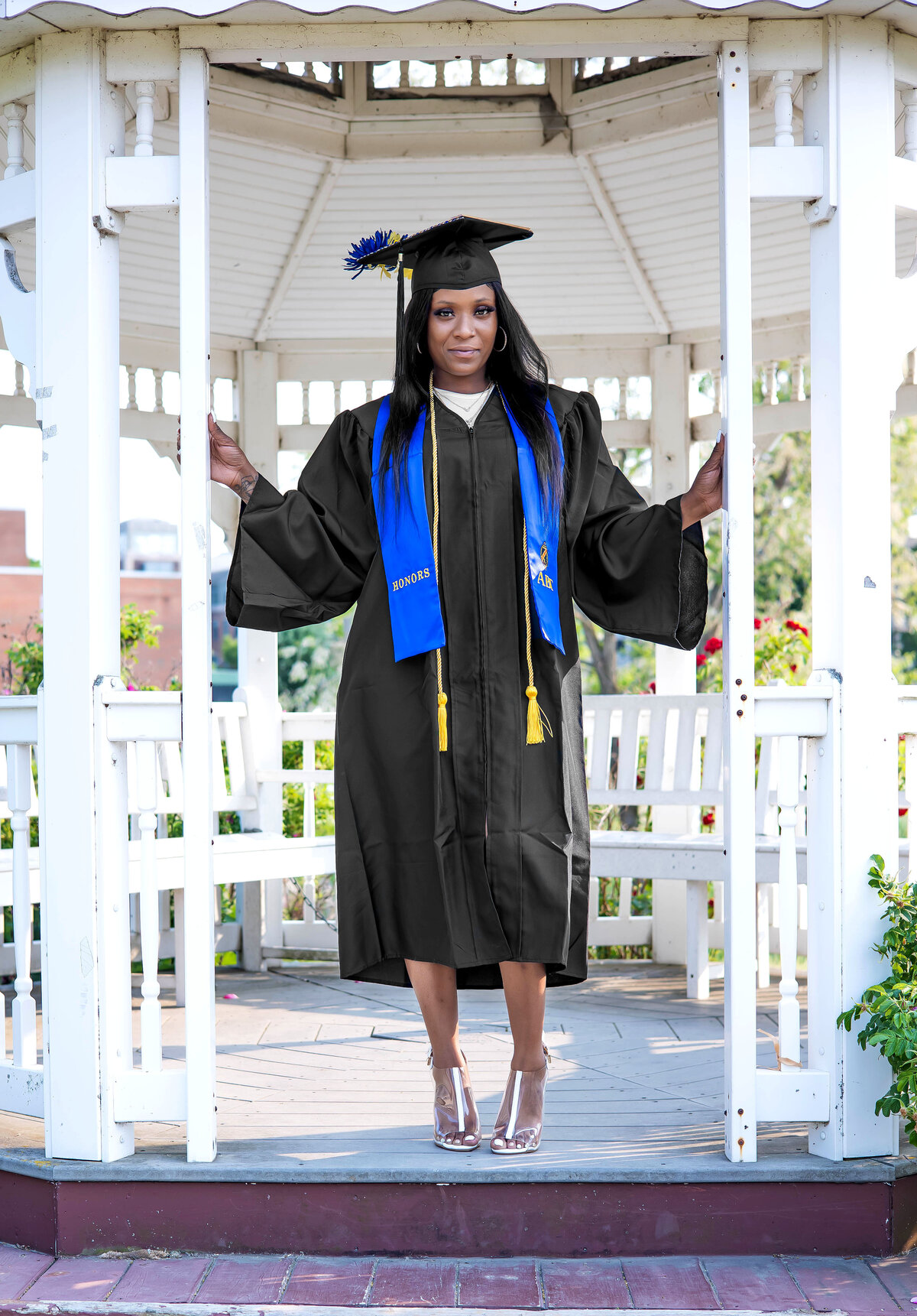 College graduate wearing black graduation cap and gown, blue honors sash, and yellow cord in a gazebo