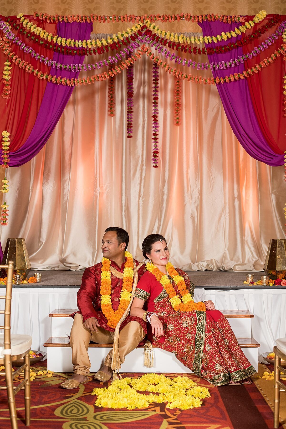 Hindu groom and bride at an Indian wedding in Nashville sitting on the steps of the peach, magenta and red mandap. The bride is wearing a red, green and gold saree and the groom is wearing a gold and red sherwani. The Indian Bride and groom are wearing orange and yellow varmalas with yellow flower petals on the red carpet at Embassy Suites, Nashville