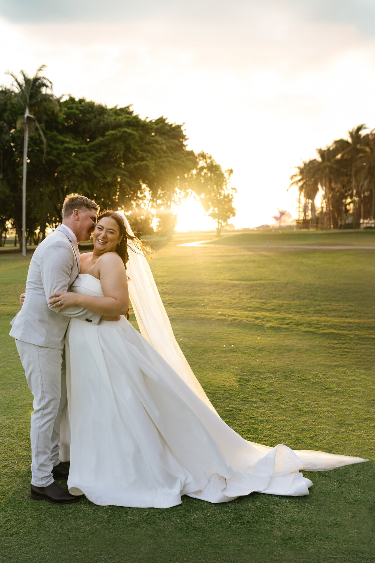 The bride giggles as the groom whispers into her ear, the sun setting behind them. 